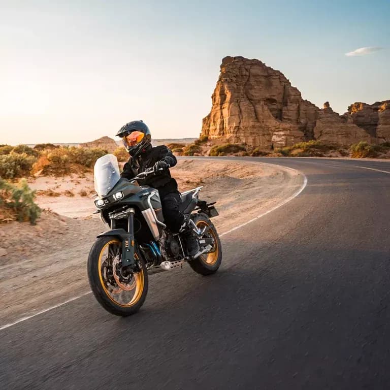 Piloto con casco y equipo negro conduciendo una motocicleta KOVE adventure gris por una carretera curva en un paisaje desértico de roca al atardecer