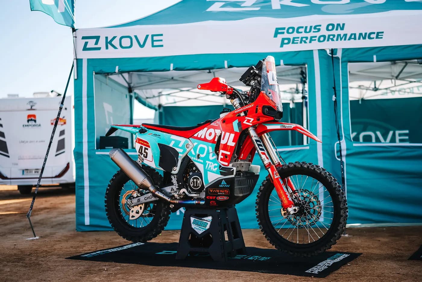 Motocicleta KOVE de rally sobre un caballete bajo una carpa del equipo KOVE con el lema «Focus on Performance»; dorsal 45 y el nombre «SUNIER» legibles