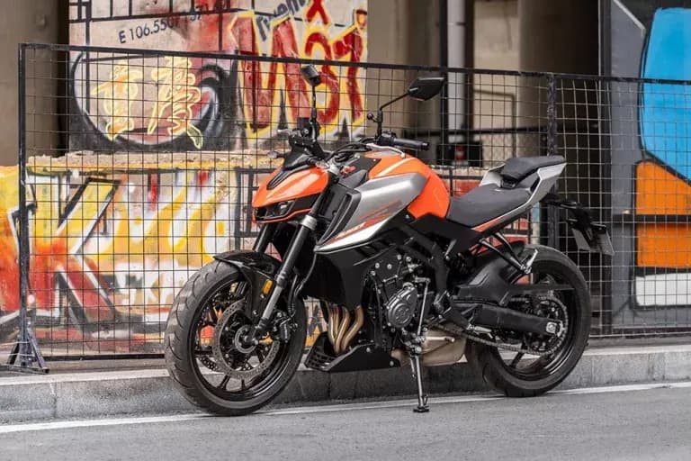 Motocicleta KOVE naked naranja y plata estacionada en la calle frente a una valla metálica y un muro cubierto de grafiti