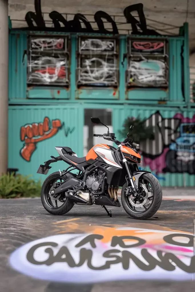 Motocicleta KOVE naked naranja, blanca y negra frente a contenedores turquesa cubiertos de grafiti
