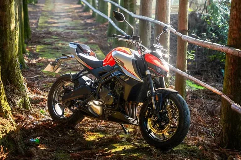 Motocicleta KOVE naked plata, roja y negra con llantas doradas, estacionada en un sendero de bosque de coníferas