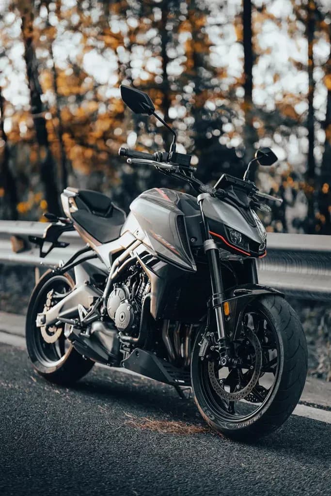 Motocicleta KOVE naked gris y negra junto a un guardarraíl de carretera con follaje otoñal desenfocado detrás