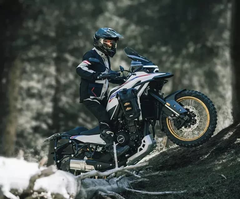 Piloto con casco y chaqueta de enduro conduciendo una motocicleta KOVE adventure blanca y azul por un sendero de tierra en un bosque